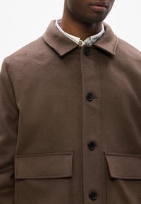 Cappotto in misto lana marrone con colletto, bottoni frontali e due ampie tasche a patta. Presenta una texture morbida e un design snello.