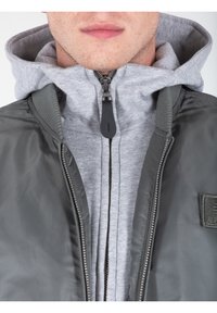 Giacca bomber grigia dal finish liscio, con colletto alto, chiusura zip anteriore e un cappuccio grigio sovrapposto. Inserti in maglia strutturata.