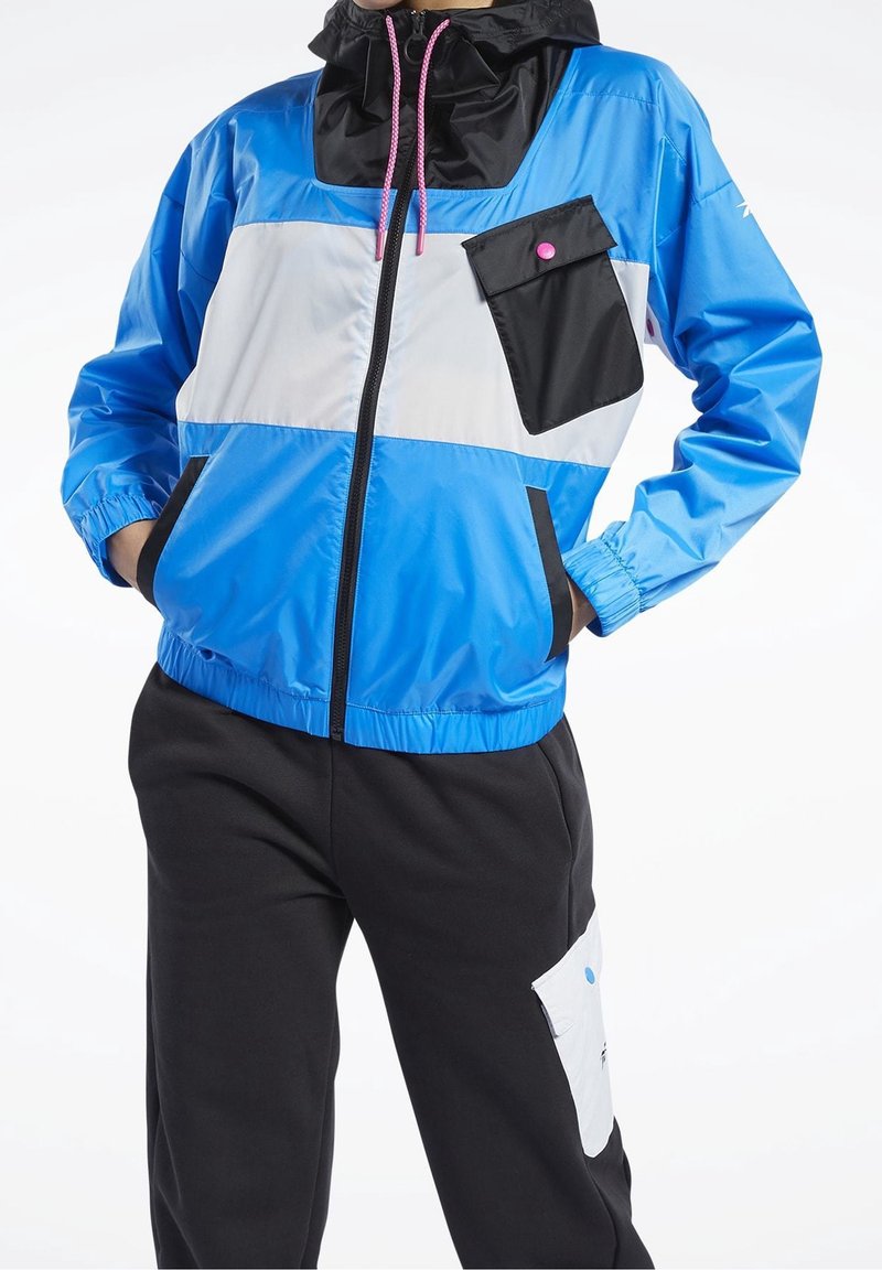 Personne portant une veste zippée bleue, blanche et noire avec des cordons roses et un pantalon noir avec une poche blanche sur la cuisse.