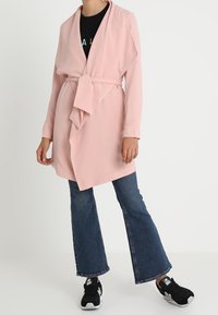 Cappotto avvolgente rosa chiaro con cintura e maniche lunghe, davanti drappeggiato, abbinato a jeans svasati scuri e sneakers nere. Tessuto dalla texture liscia.