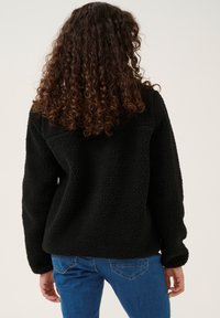 Svart fleecejacka med texturerad yta, långa ärmar och avslappnad passform, bärs över blå jeans. Slät tygbaksida visas.