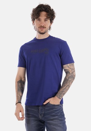 Uomo con capelli ricci che indossa una t-shirt blu Armani Exchange e jeans, mostrando tatuaggi dettagliati su entrambe le braccia, in piedi su sfondo bianco.