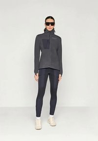 Naine, kes kannab musta lukuga jope, musti retuuse, millel on plaastrid, valgeid tossusid ja valgeid sokke, seisab valge tausta ees suured mustad päikeseprillid peas.