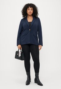 Blazer marine à fermeture à un bouton et deux poches avant, associé à des leggings noirs et des bottines, tenant un sac à main noir.