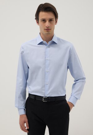 Mann trägt ein hellblau gestreiftes Hemd, in schwarze Hose mit schwarzem Gürtel gesteckt, steht vor einem einfarbigen hellen Hintergrund.