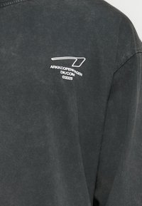 Svart kortermet skjorte laget av myk bomull med en falmet tekstur. Har et hvitt logotrykk på øvre venstre side.