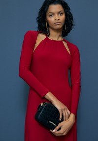 Červené šaty s dlouhým rukávem, staženým výstřihem a vykrojenými rameny, doplněné černou prošívanou kabelkou se zlatými kovovými doplňky.