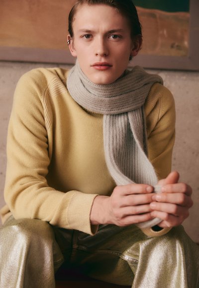 Gelber Strickpullover, hellgrauer gerippter Schal, metallisch silberne Hose. Nahaufnahme von Händen, die den Schal halten, neutraler Hintergrund mit strukturierten Wänden.