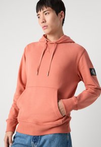 Felpa rosa con cappuccio, tasca a marsupio, polsini a costine e patch con logo sulla manica. Tessuto morbido con una consistenza liscia.