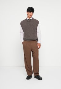 Pull sans manches en maille marron sur une chemise blanche, associé à un pantalon large marron et des chaussures noires. Tissu texturé, coupe décontractée.