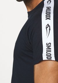 Marinblå t-shirt med kort ärm och vita sidostråk, som har svart text och en grafisk design. Mjuk bomullstyg med sömdetaljer.