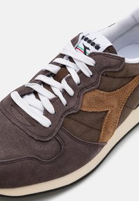 Zapatillas de ante y tela marrón con cordones blancos, que presentan un acento de ante color beige y una superficie texturizada. La suela de goma completa el diseño.