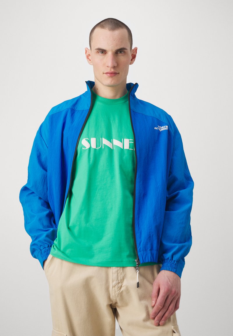 Giacca leggera blu con colletto alto, chiusura con cerniera e polsini elasticizzati sopra una maglietta verde con testo bianco. Pantaloni beige.