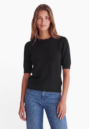 Femme aux longs cheveux bruns portant un pull noir à manches courtes et un jean bleu, debout devant un fond blanc uni.