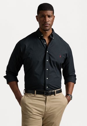 Camicia nera con bottoni e maniche lunghe, caratterizzata da un piccolo logo rosso. Abbinata a pantaloni color khaki e una cintura nera. Texture del tessuto liscia.