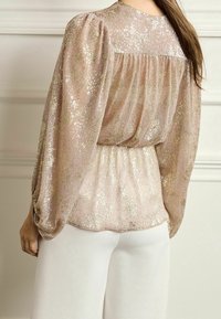 Blouse rose pâle à sequins avec manches bouffantes et taille froncée, présentant une texture lisse et des accents métalliques subtils. Associée à un pantalon blanc.