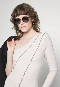 Top bege claro com textura em canalé, com decote ombro a descoberto e um detalhe ondulado castanho. Óculos de sol redondos e escuros, e brincos de argola verdes complementam o visual.