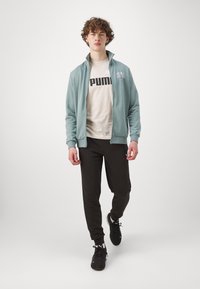 Jasnoniebieska kurtka zapinana na suwak nałożona na jasno kremowy t-shirt z dużym czarnym logo, w połączeniu z czarnymi spodniami i czarnymi trampkami. Gładka tkanina i luźny krój.