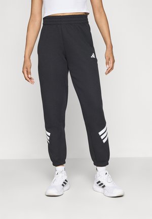 Femme portant un pantalon de survêtement Adidas noir avec des bandes blanches et des baskets Adidas blanches sur un fond uni.