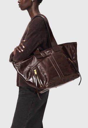 Persoon met over elkaar geslagen armen, gekleed in een donkere trui en spijkerbroek, met een grote glanzende donkerbruine leren shopper met ritszakken aan de voorkant.