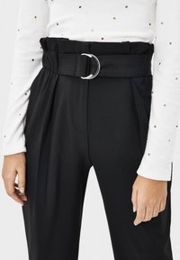 Schwarze hoch taillierte Hose mit einem breiten Gürtel, der einen silbernen Metallring hat. Der Stoff ist glatt mit Falten und einer maßgeschneiderten Passform.