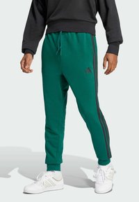 Groene katoenen joggingbroek met twee zwarte zijstrepen, elastische tailleband met trekkoord en aansluitende boorden. Zwart logo op de dij.