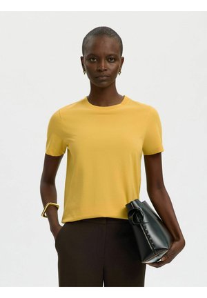 Kvinde iført en gul kortærmet skjorte og mørke bukser, der holder en sort foldet clutchpose, mens den ene hånd er i lommen.