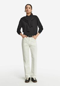 Schwarzes Langarmoberteil mit einem gebundenen Detail am Hals, kombiniert mit weißen geraden Jeans und schwarzen Sneakers mit beigen Schnürsenkeln.