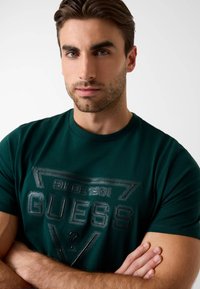 Zielona bawełniana koszulka z wyeksponowanym czarnym logo "GUESS" oraz napisem "SINCE 1981" na środku. Krótkie rękawy, okrągły dekolt, gładka faktura.