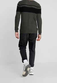 Dunkelgrüner Strickpullover mit horizontalen schwarzen und grauen Streifen, kombiniert mit schwarzen, schmal geschnittenen Hosen und weißen Socken, ergänzt durch Sneakers.