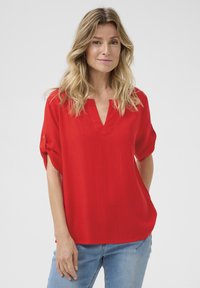 Blusa rossa a maniche corte con scollatura a V, realizzata in un tessuto leggero. Presenta maniche arrotolate e una vestibilità comoda, abbinata a jeans blu.