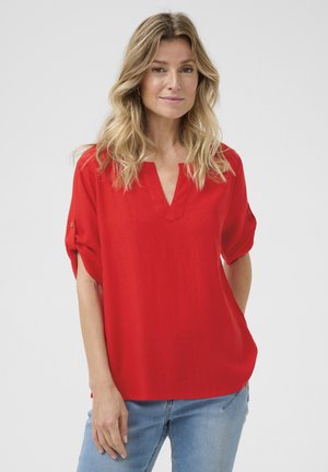 Röd kortärmad blus med V-ringning, tillverkad av ett lätt material. Har rullade ärmar och en avslappnad passform, kombinerad med blå jeans.