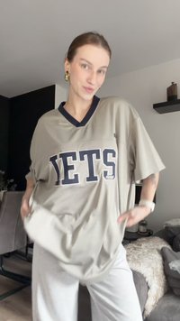 Šviesiai pilkas džersis su užrašu "JETS" tamsiai mėlyna spalva. V-kaklo dizainas, laisvas siluetas, trumpi rankoviai, tekstūruota medžiaga, derinama su šviesių spalvų kelnėmis.