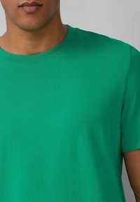 Zielona bawełniana koszulka z okrągłym dekoltem i krótkimi rękawami. Posiada subtelne wytłoczone logo na piersi. Gładka tekstura, luźny krój.