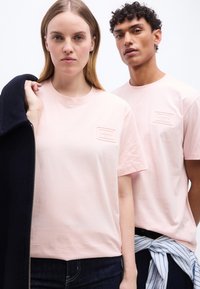 Δύο μοντέλα στέκονται δίπλα-δίπλα φορώντας ανοιχτό ροζ μπλουζάκια Lacoste· η γυναίκα κρατάει ένα σκούρο σακάκι στον ώμο της.