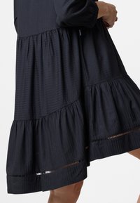 Schwarzes Kleid mit einem gestuften Design, gerippter Textur und transparenten horizontalen Akzenten am Saum. Lange Ärmel mit gerafften Manschetten.