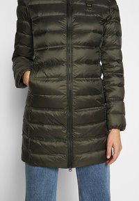 Dark green padded coat with a zip front, featuring horizontal quilted sections and a high collar. Sleeves are puffed and fitted.