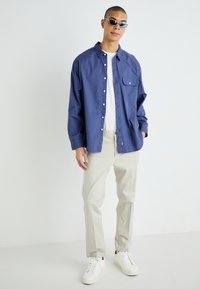 Camisa de botones ligera en color azul marino con un bolsillo en el pecho, combinada con pantalones beige claros y zapatillas blancas. Además, incluye gafas de sol azules.
