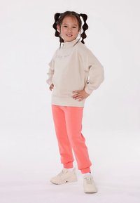 Šviesiai smėlio spalvos džemperis su siuvinėjimu, derinamas su ryškiai koralinių spalvų laisvalaikio kelnėmis ir baltais sportbačiais. Turi laisvo kirpimo stilių ir ribotas rankogalius.