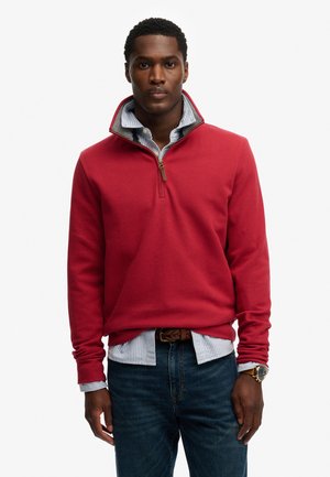 Homme portant un sweat-shirt rouge à fermeture éclair sur une chemise rayée bleue, jean foncé et une montre, debout devant un fond blanc.