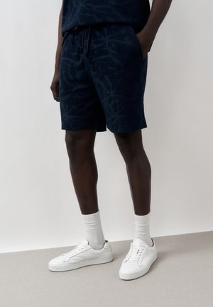 Mand iført mørke mønstrede shorts, hvide crew-sokker og hvide lavtop-sneakers, stående mod en ensfarvet lys baggrund.