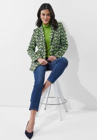 Een blazer met een groen bladpatroon over een groene col trui, gecombineerd met blauwe jeans en marineblauwe hakken, zittend op een witte kruk tegen een witte achtergrond.