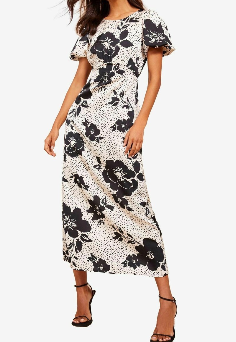 Béžové květinové maxi šaty s velkými černými květinami a puntíky, s krátkými nabíranými rukávy a upnutým pasem. Černé sandály na tenkých páscích.