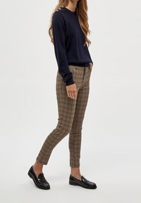 Marinblå tröja med knäppdetaljer på axeln, tillsammans med bruna rutiga byxor och svarta loafers. Tätt sittande design, mjuk textur.