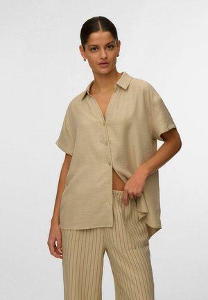 Femme aux cheveux foncés portant une chemise beige à manches courtes boutonnée et un pantalon beige à fines rayures verticales, tenant l'ourlet de la chemise d'une main.