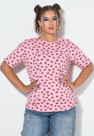 Vrouw met gevlochten haar en blauwe oogmake-up draagt een roze shirt met rood lippenpatroon en gescheurde blauwe jeans, handen in de heupen.
