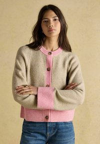 Žena s dlhými tmavými vlasmi, oblečená v béžovom a ružovom zapínacom kardigáne a modrých džínsoch, so založenými ramenami na neutrálnom pozadí.