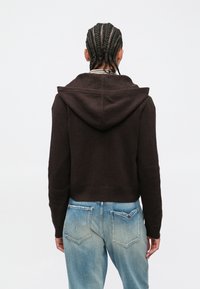 Άτομο με πλεξούδες, που φοράει σκούρο καφέ πουλόβερ με κουκούλα και ανοιχτό μπλε ξεθωριασμένο τζιν, στέκεται με την πλάτη γυρισμένη σε απλό φόντο.