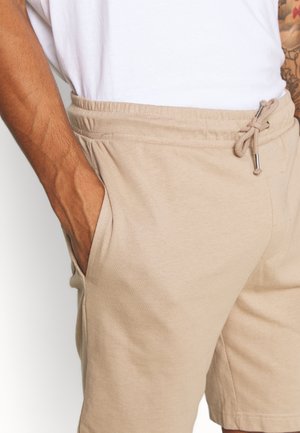 Mann trägt beige Shorts mit Kordelzug und ein weißes Hemd, linke Hand in der Shorts-Tasche, sichtbare Arm-Tattoos.