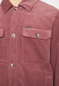 Een corduroy shirt in stofroze, met verticale ribbels, zilveren knopen en twee borstzakken met logo-label op de linkerzak.